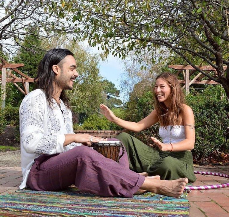 Two friends sharing a djembe drum on a rug in the park