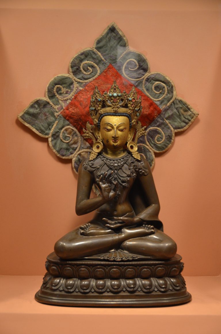 Bronze crowned Buddha seated in meditation against a terracotta wall