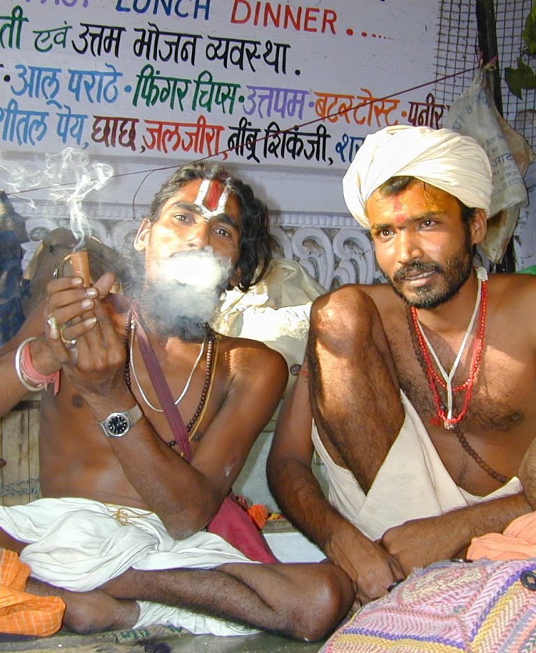 Two sadhus sharing a chillum beneath hand-painted signage