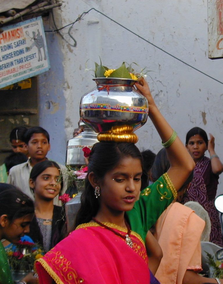 Girl balancing a brass water pot crowned with flowers on her head