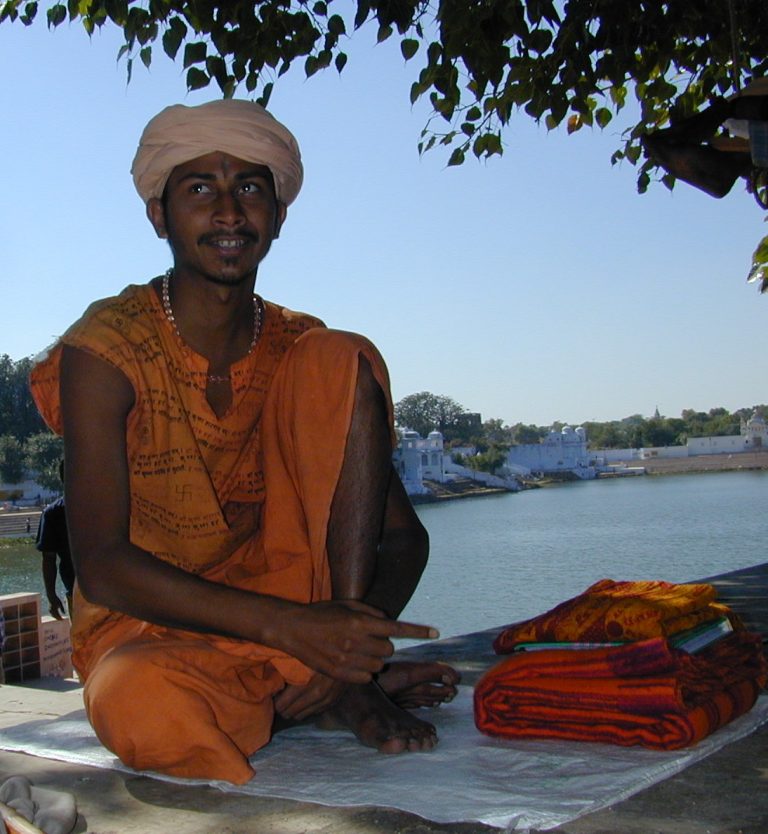 Young sadhu in orange beside folded textiles at the lake