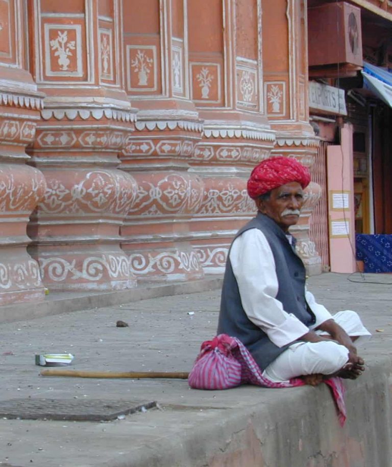 Elder in a red turban resting his cane by a carved pink wall